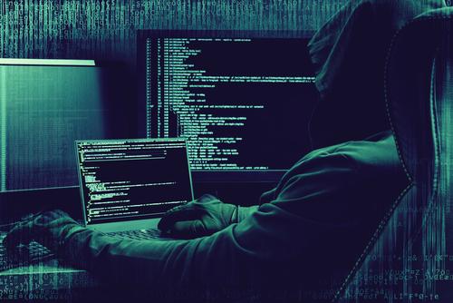 A man in a hoody sitting in front of several computer screens apparently hacking data