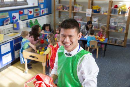 A male teacher in a pre-school classroom. He is holding some aprons