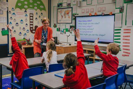 The Covid learning gap is starting to narrow. Female teacher is teaching shapes to her primary school students. She is asking hem a question and some of the students have their hand in the air to answer.